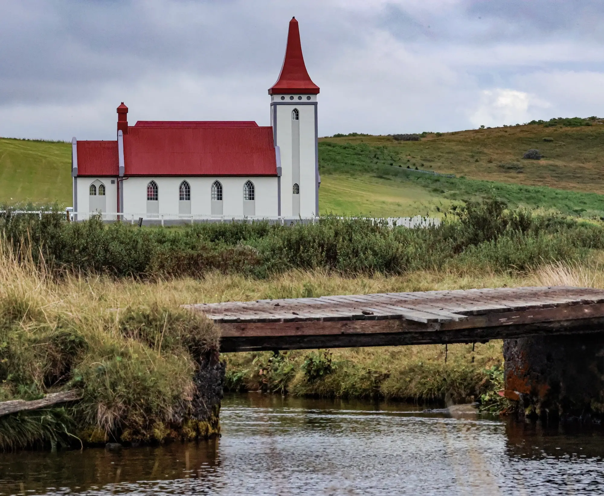 Eine weiße Kirche mit rotem Dach steht hinter einer Holzbrücke über einen Bach in einem grasbewachsenen Feld.