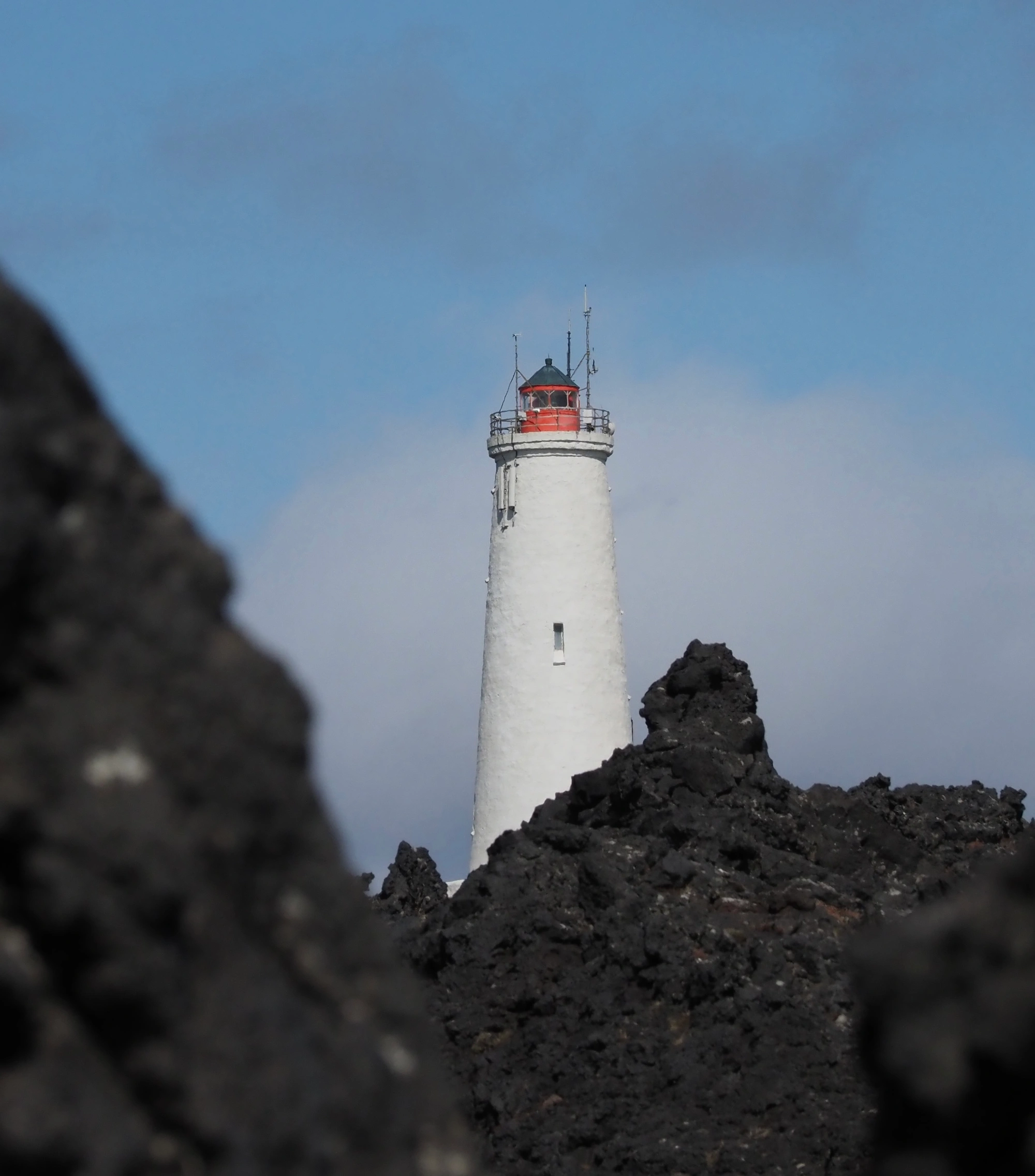 Auf der Insel erhebt sich hinter dunklen Vulkanfelsen und blauem Himmel ein weißer Leuchtturm mit einer roten Spitze.