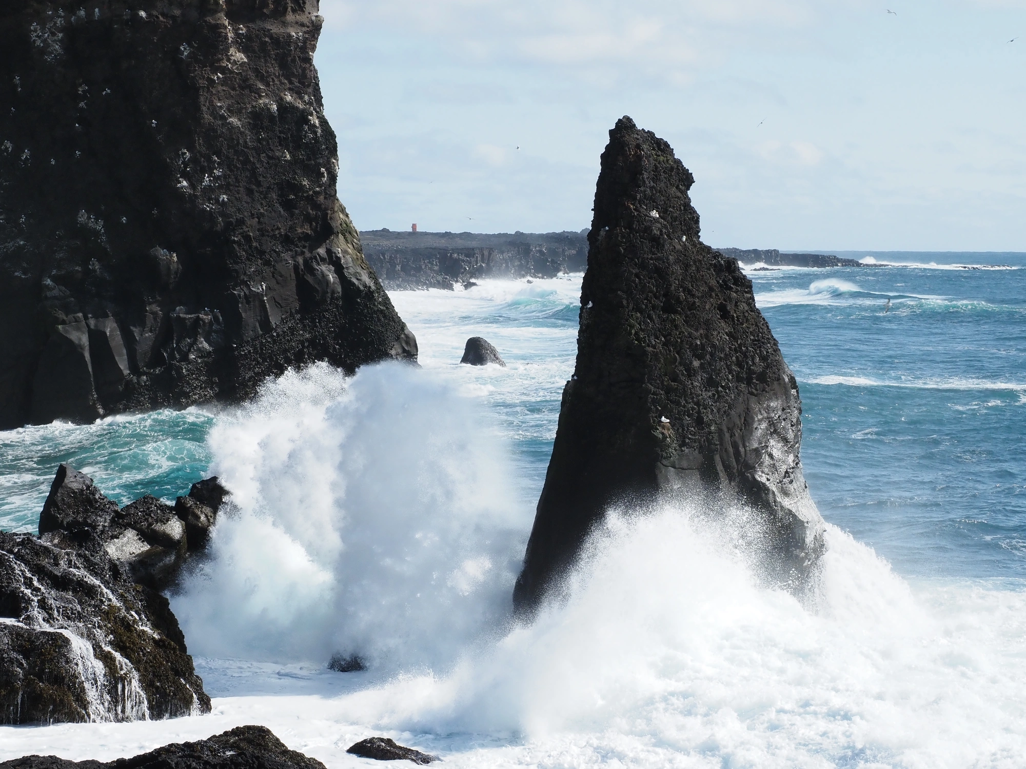 An einem hellen, teilweise bewölkten Tag schlagen die Wellen gegen die zerklüfteten schwarzen Felsen am Meer der Insel.