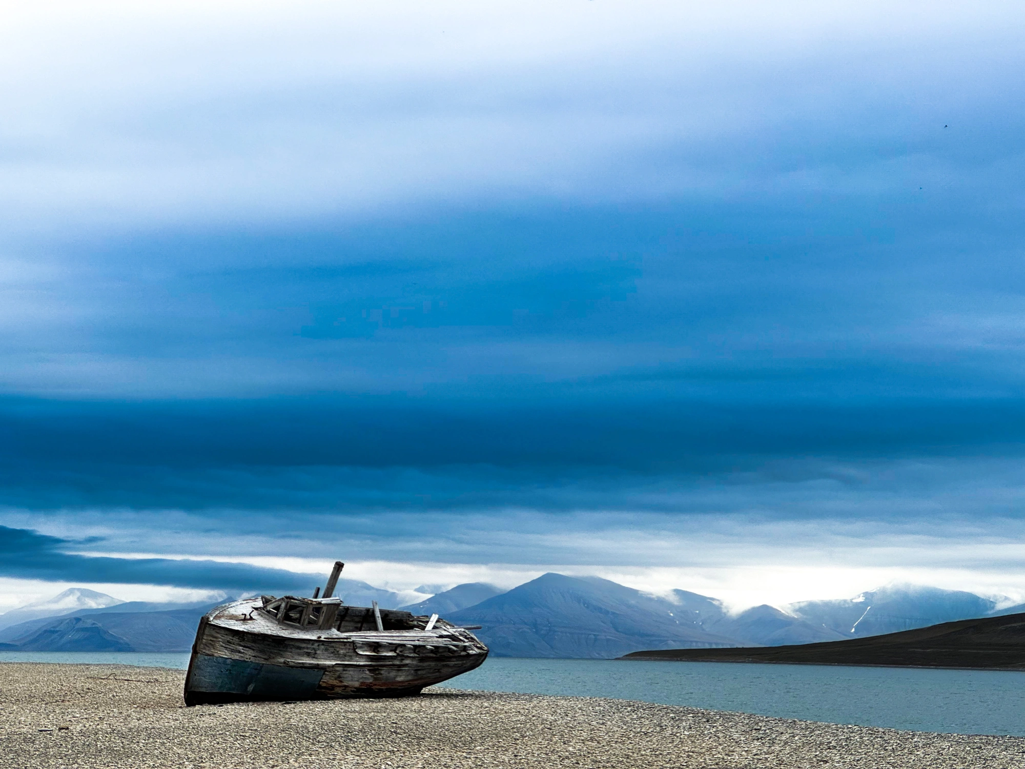Ein altes Holzboot liegt auf einem Kieselstrand in Spitzbergen mit Bergen und bewölktem Himmel im Hintergrund.