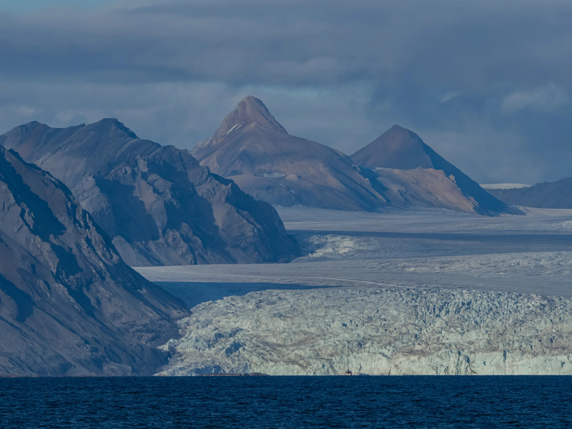 Unter einem wolkenverhangenen Himmel treffen zerklüftete Berge und ein Gletscher auf den Ozean von Reviere.