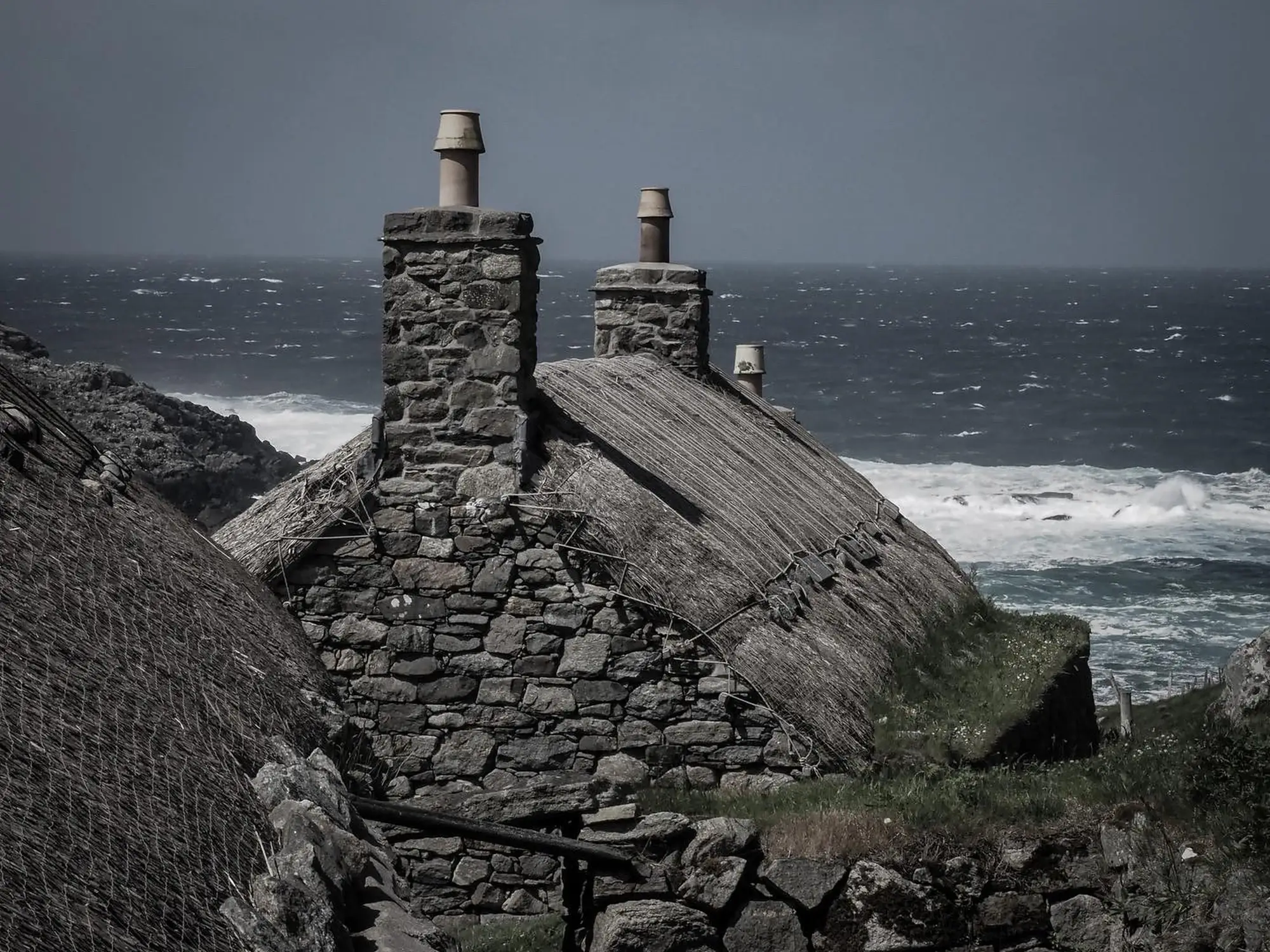 Steinhäuser mit Strohdächern säumen die Küste der Hebriden unter einem wolkenverhangenen, felsigen Küstenhimmel.
