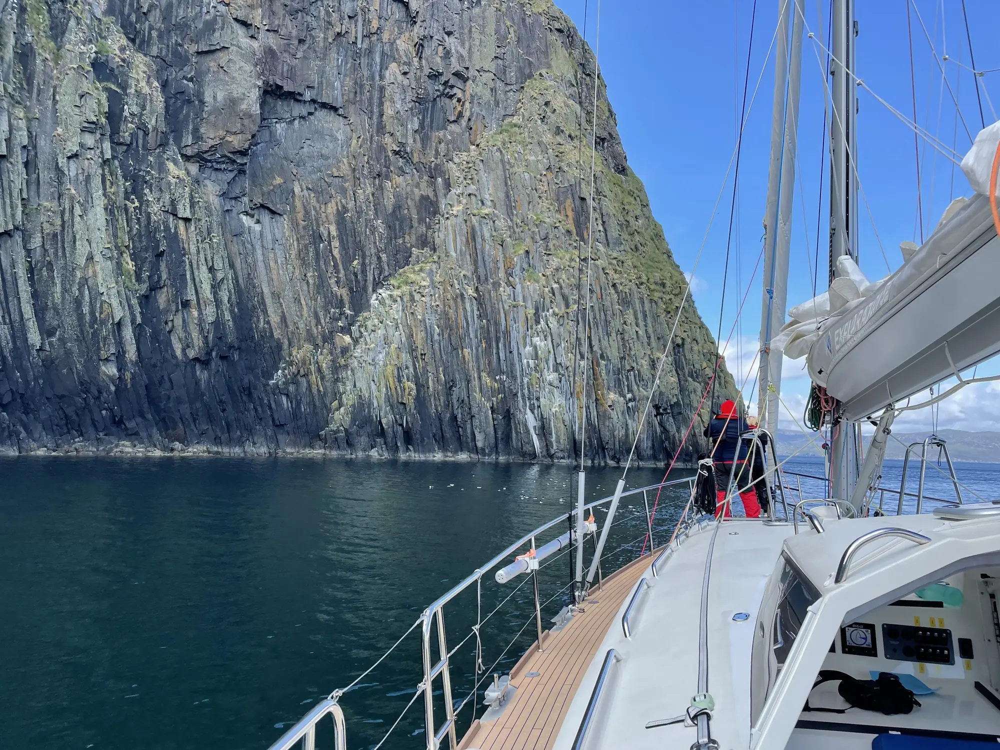 Eine Person auf einem Segelboot nähert sich den steilen, felsigen Hebriden-Klippen über ruhigem, dunklem Wasser.
