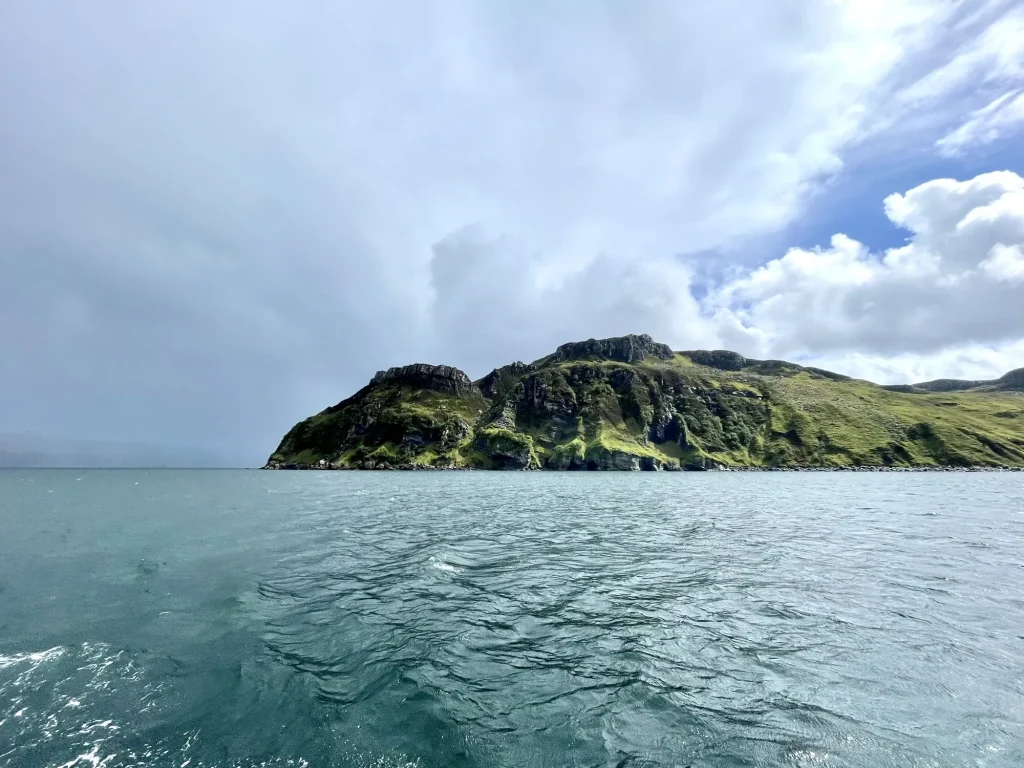 Eine felsige, grüne Hebrideninsel unter einem teilweise bewölkten Himmel, umgeben von kabbeligem, blauem Wasser.