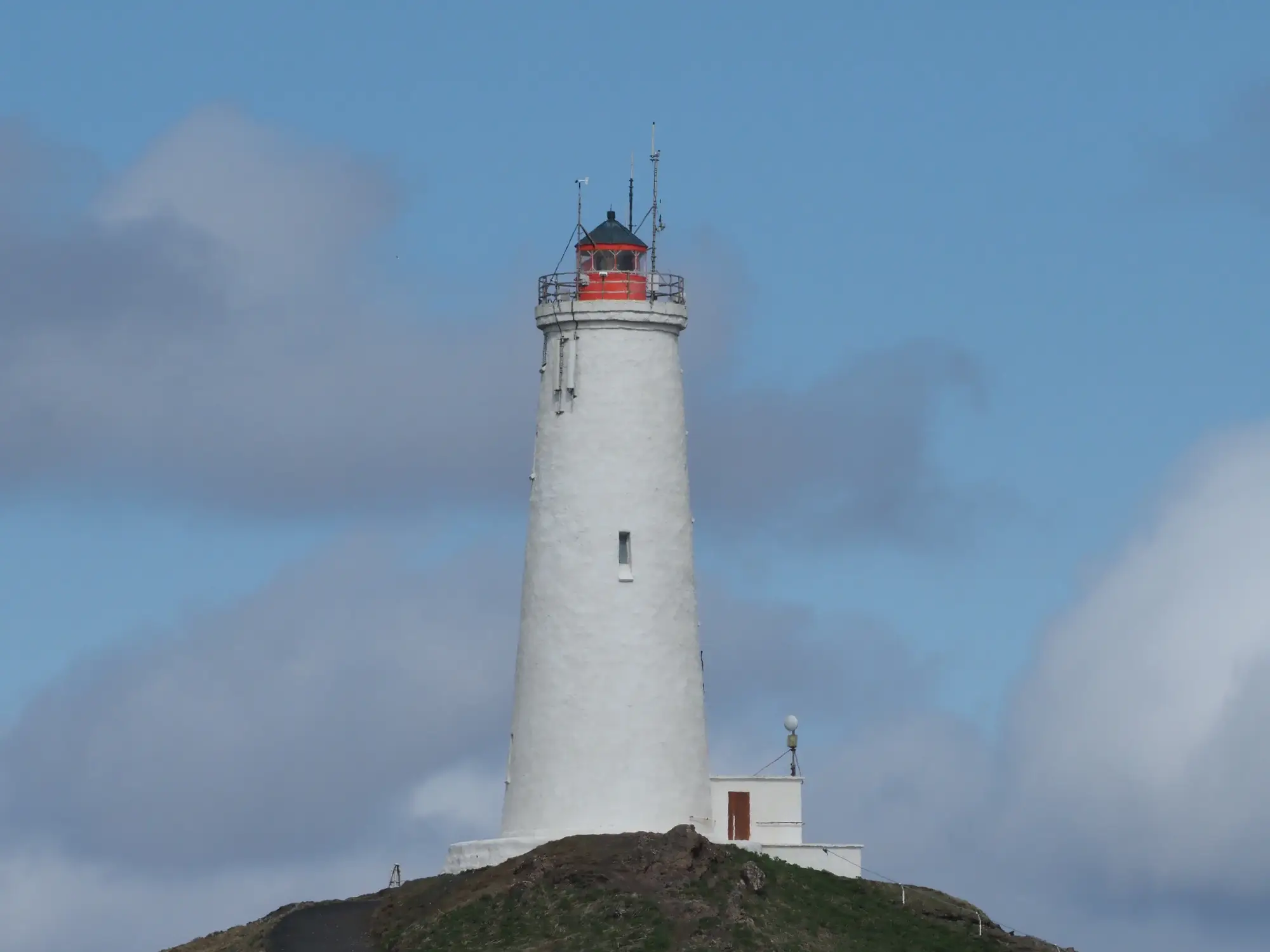 Ein weißer Leuchtturm mit roter Spitze steht auf einem Hebridenhügel unter einem blauen Himmel mit Wolken.