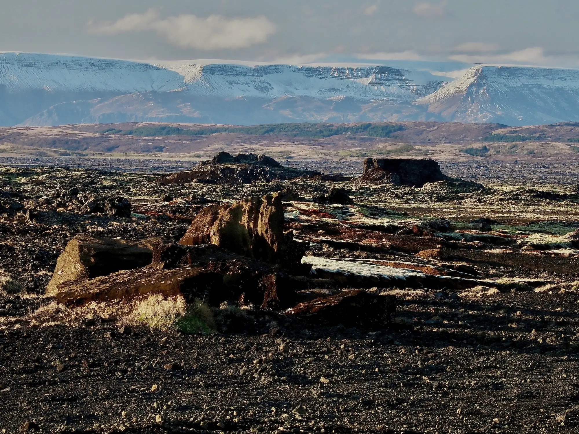 Felsige Vulkanebene in den Hebriden mit schneebedeckten Bergen in der Ferne unter einem bewölkten Himmel.
