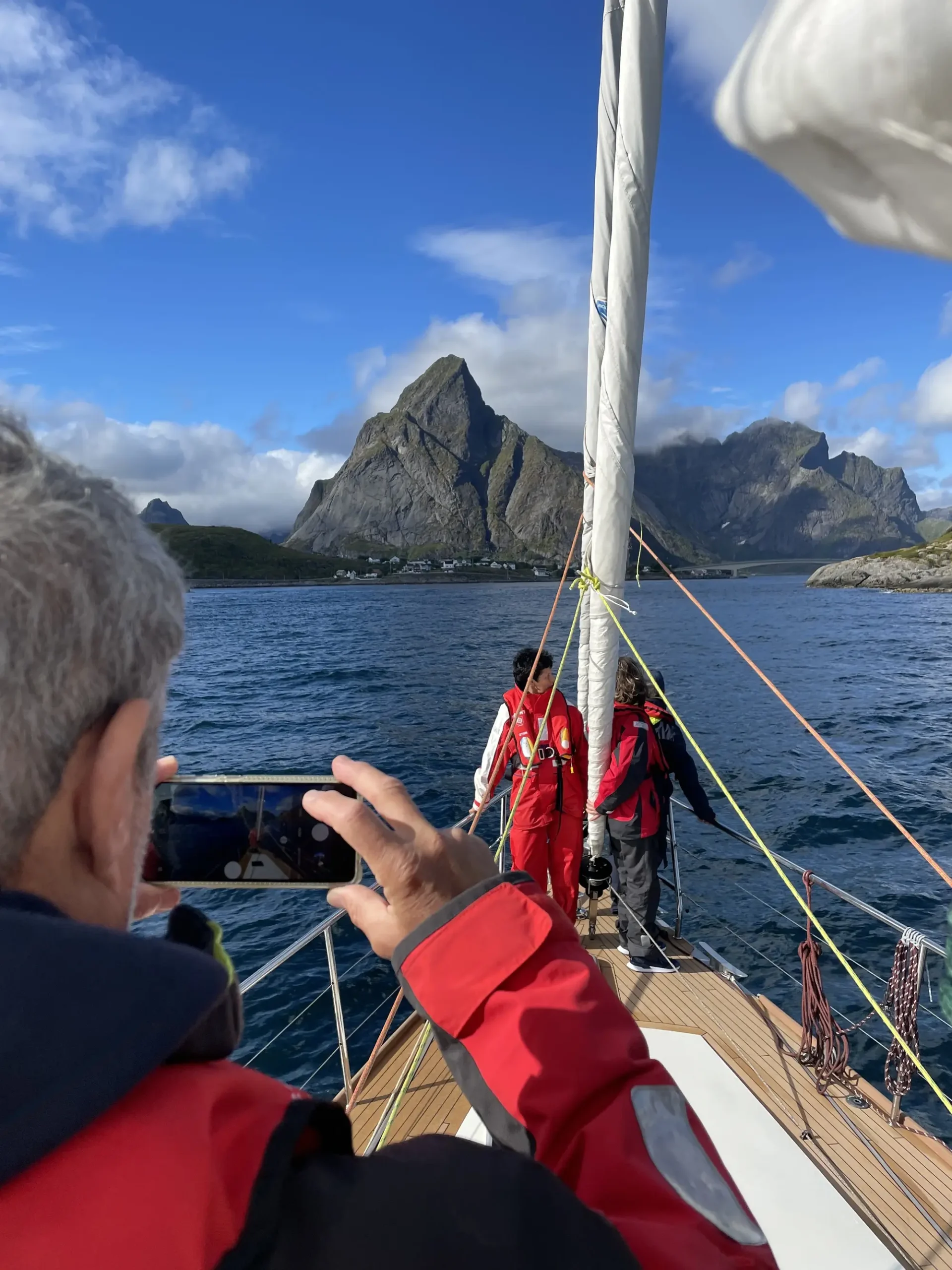 People on a sailing boat taking photos, with the Lofoten Islands and the blue sky in the background.
