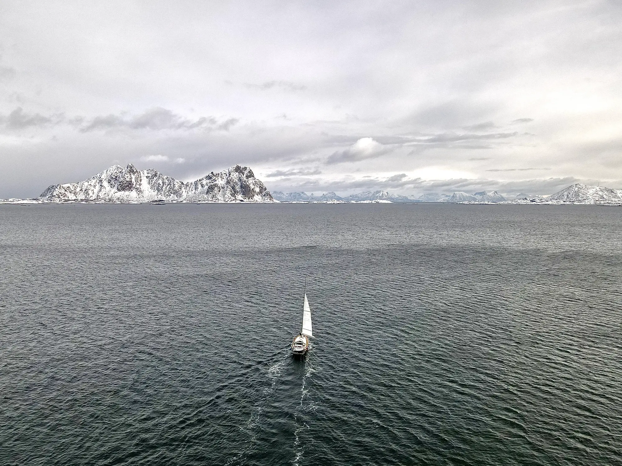 A sailing boat glides on a calm sea with the snow-capped mountains of Lofoten under a cloudy sky.