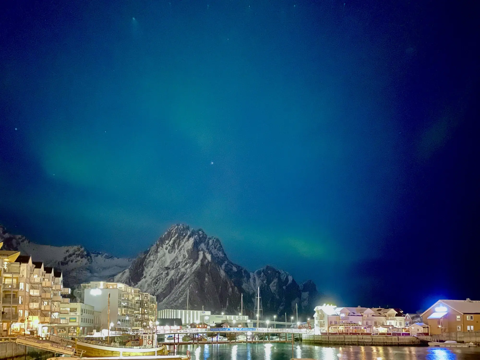 Nordlichter beleuchten die schneebedeckten Berge der Lofoten und die Gebäude am Wasser bei Nacht.