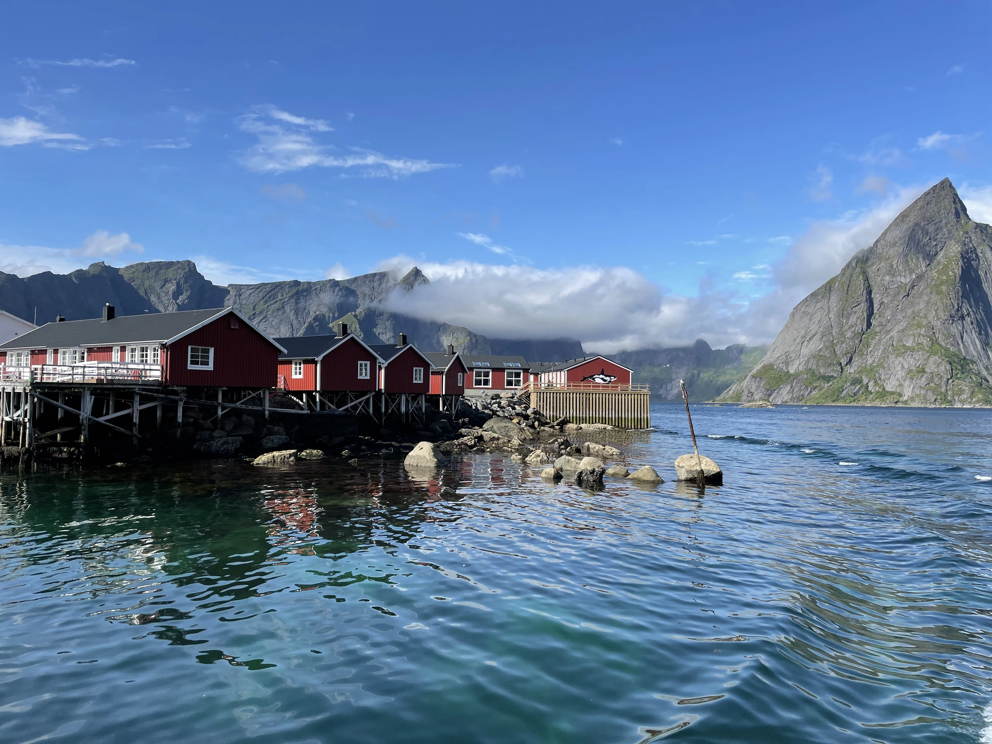 Red huts on stilts on the clear waters of Lofoten, with mountains and blue sky in the background.