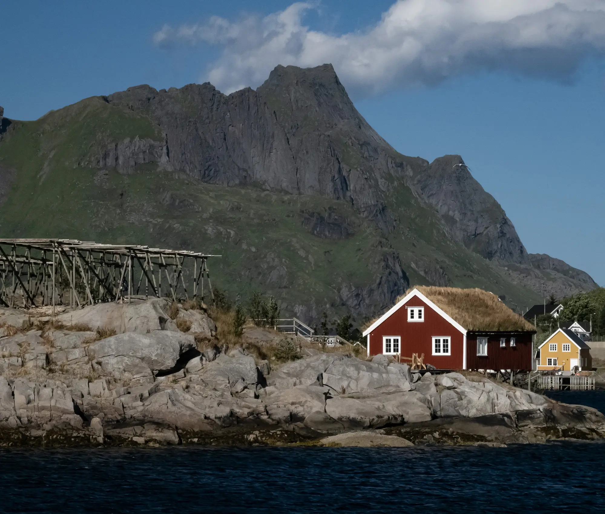 Red house with grass roof on the rocky shore of Lofoten, mountains and blue sky in the background.