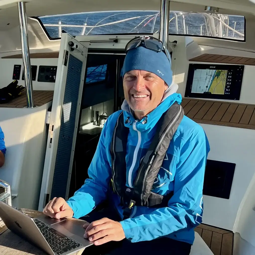 Mann in blauer Jacke lächelt und benutzt einen Laptop auf einem Segelboot während seiner Mission.