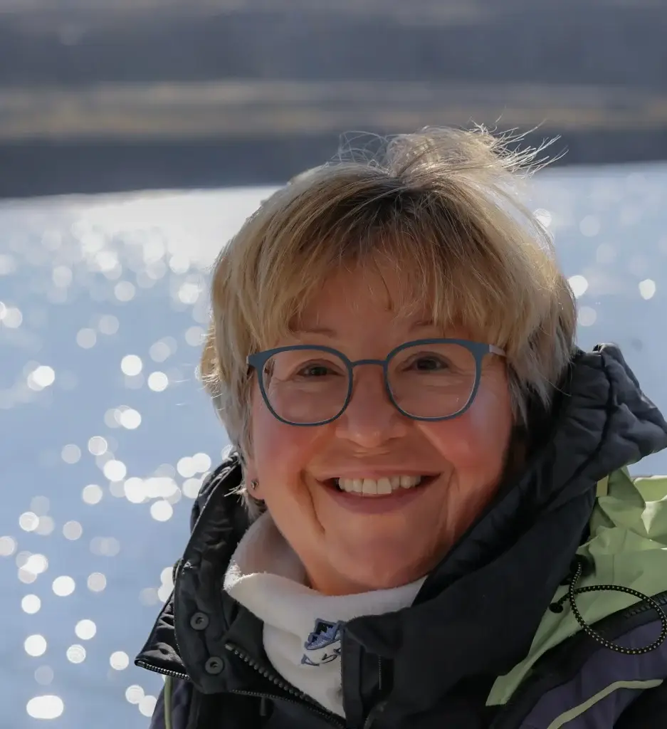 Lächelnde Frau mit Brille und Winterjacke an einem sonnenbeschienenen, glitzernden See auf einer Mission.