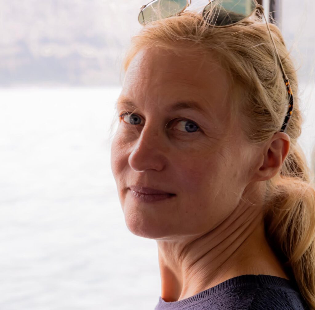 Frau mit blondem Haar und Sonnenbrille, auf einer Mission, während sie in der Nähe von Wasser über ihre Schulter schaut.