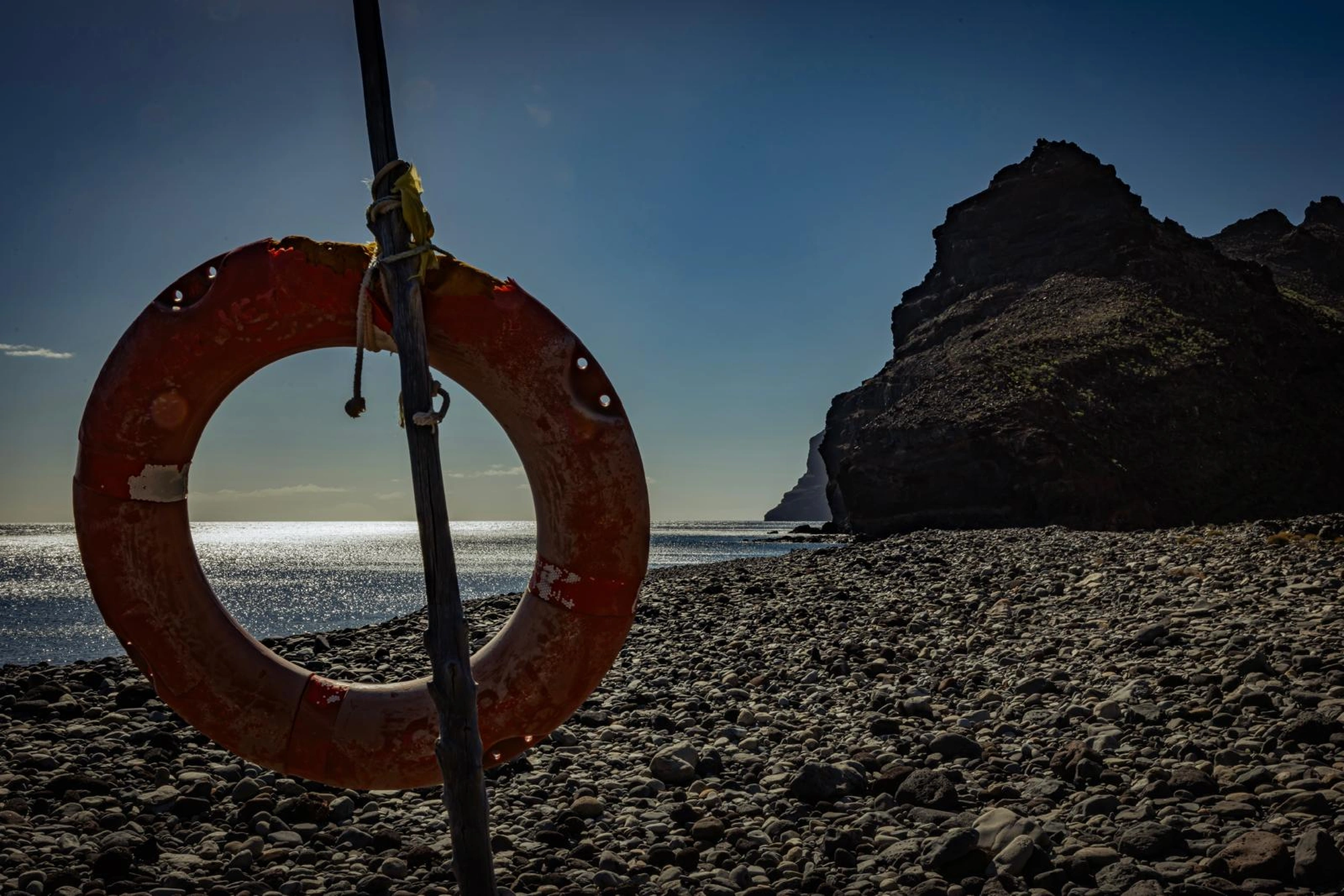 Ein Rettungsring hängt an einem felsigen Strand mit OceanLife-Kanaren Leg 1 - 261219 im Hintergrund.