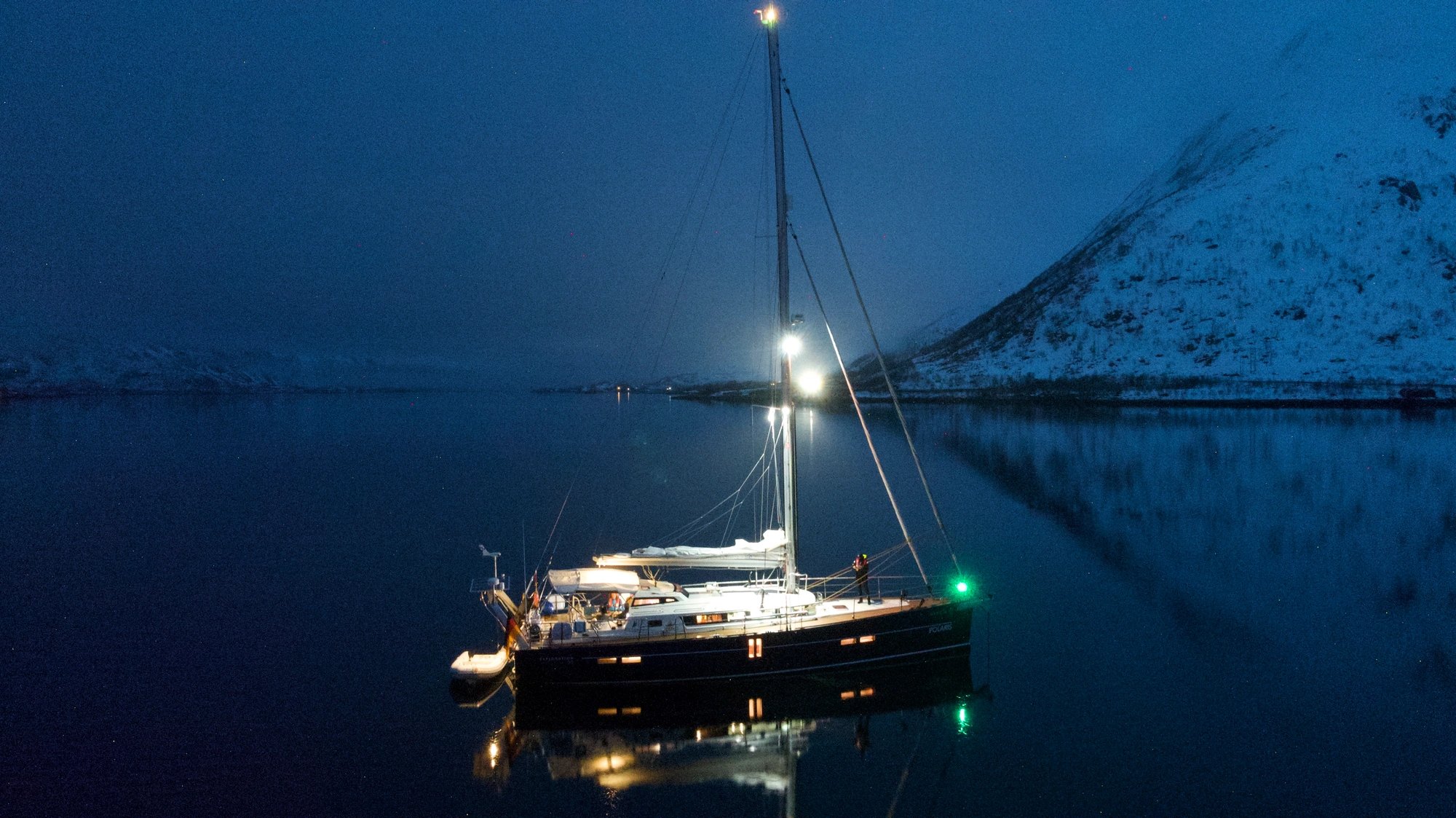 Segel & Ski Lofoten 1 - 270320: Ein Segelboot mit Lichtern schwimmt in der Abenddämmerung auf ruhigem Wasser.