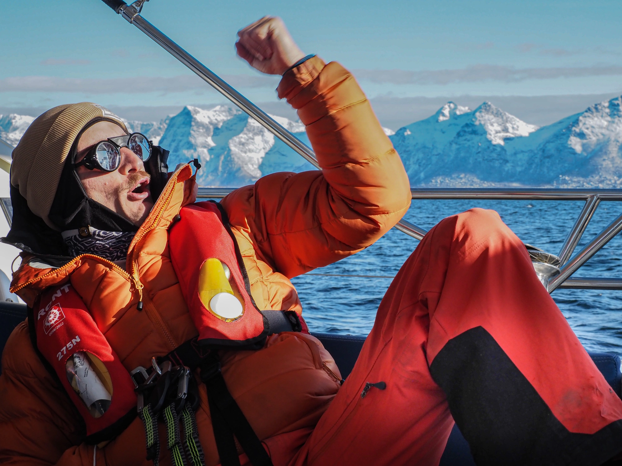 Person in orangefarbener Jacke feiert auf einem Boot mit verschneiten Lofoten im Hintergrund. Segeln & Skifahren Lofoten 1.