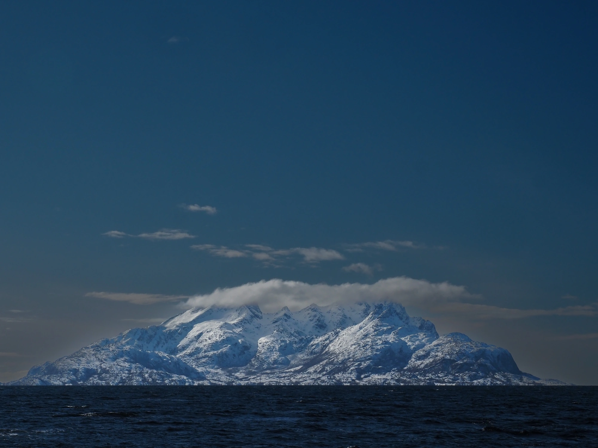 Segeln & Skifahren Lofoten 1 - 270320: Ein schneebedeckter Gipfel ragt über das dunkle Meer unter einem tiefblauen Himmel.