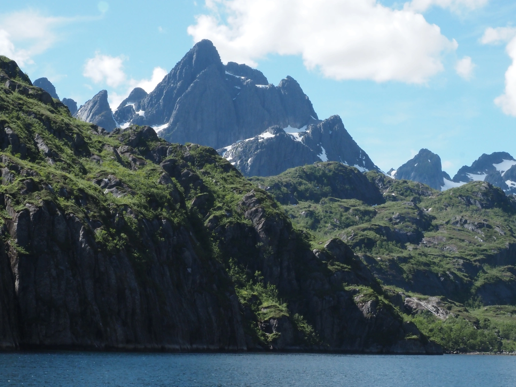 Schneebedeckte, felsige Gipfel der Lofoten ragen über einen blauen See unter den Wolken empor. Lofoten, Etappe 1 – 27.05.15.