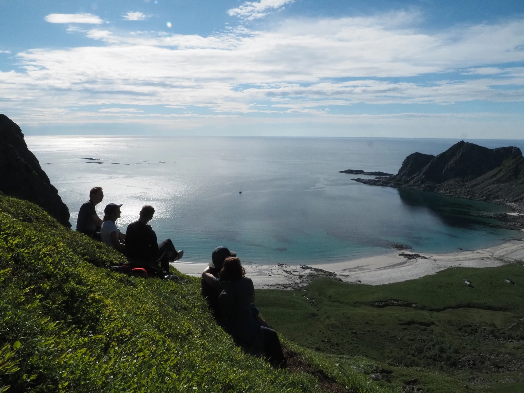 Fünf Personen genießen auf einem grasbewachsenen Hügel den Blick auf die ruhige Bucht und das Meeresleben während der ersten Etappe der Lofoten-Reise – 27.05.15.